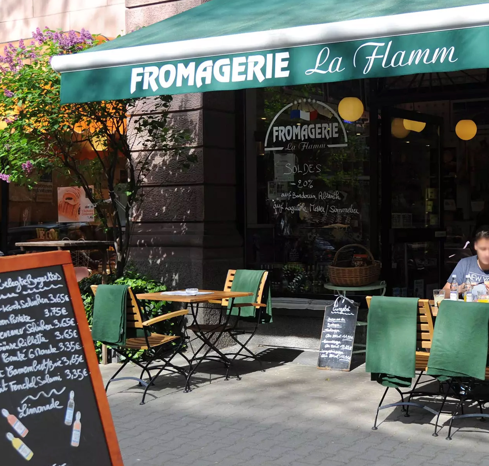 Einladendes Café 'Fromagerie La Flamm' mit Außenbereich, Tischen und Stühlen, umgeben von grünen Pflanzen.