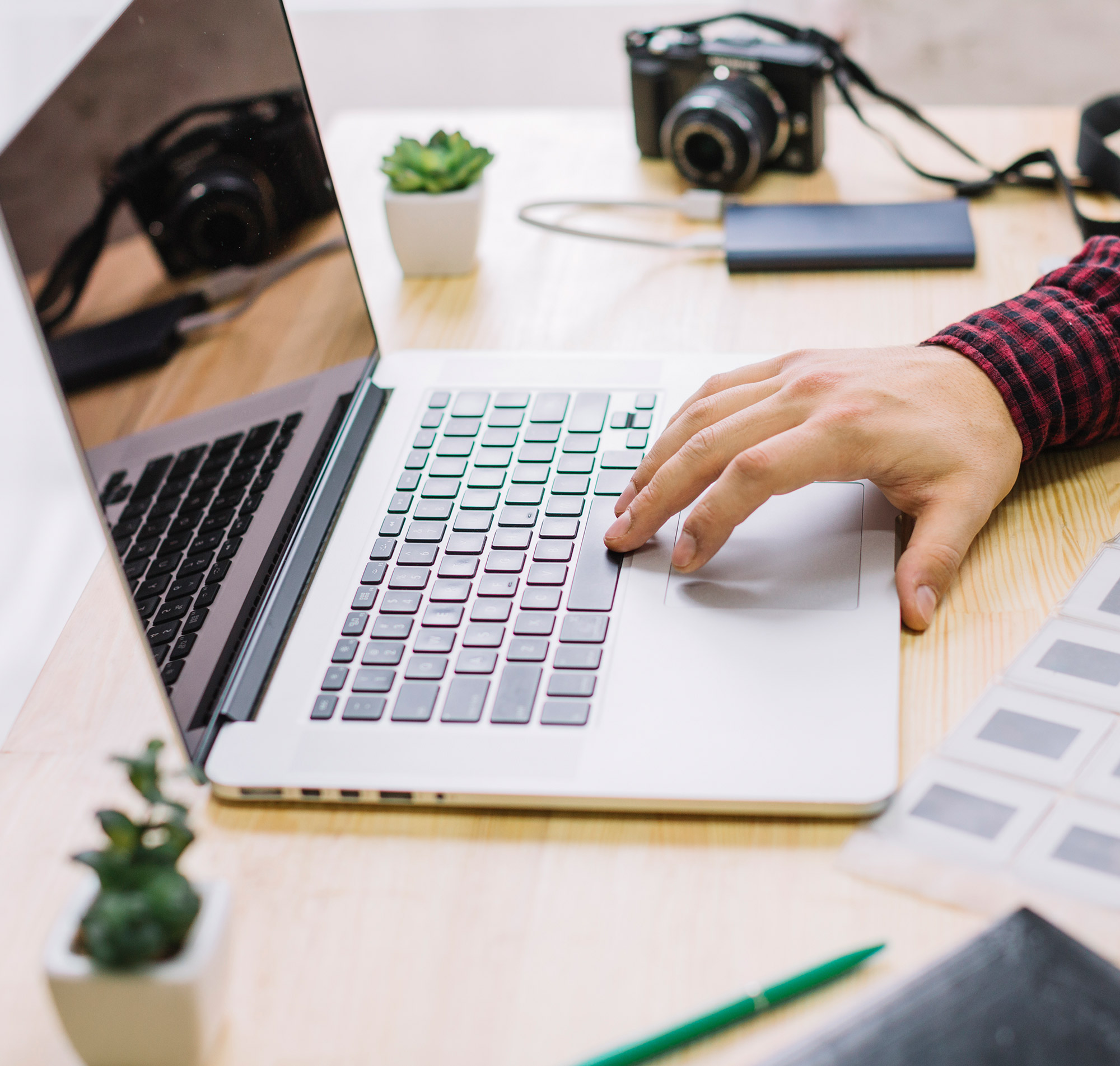 Eine Hand tippt auf einer Laptop-Tastatur, umgeben von Pflanzen und Kameras auf einem Holztisch.