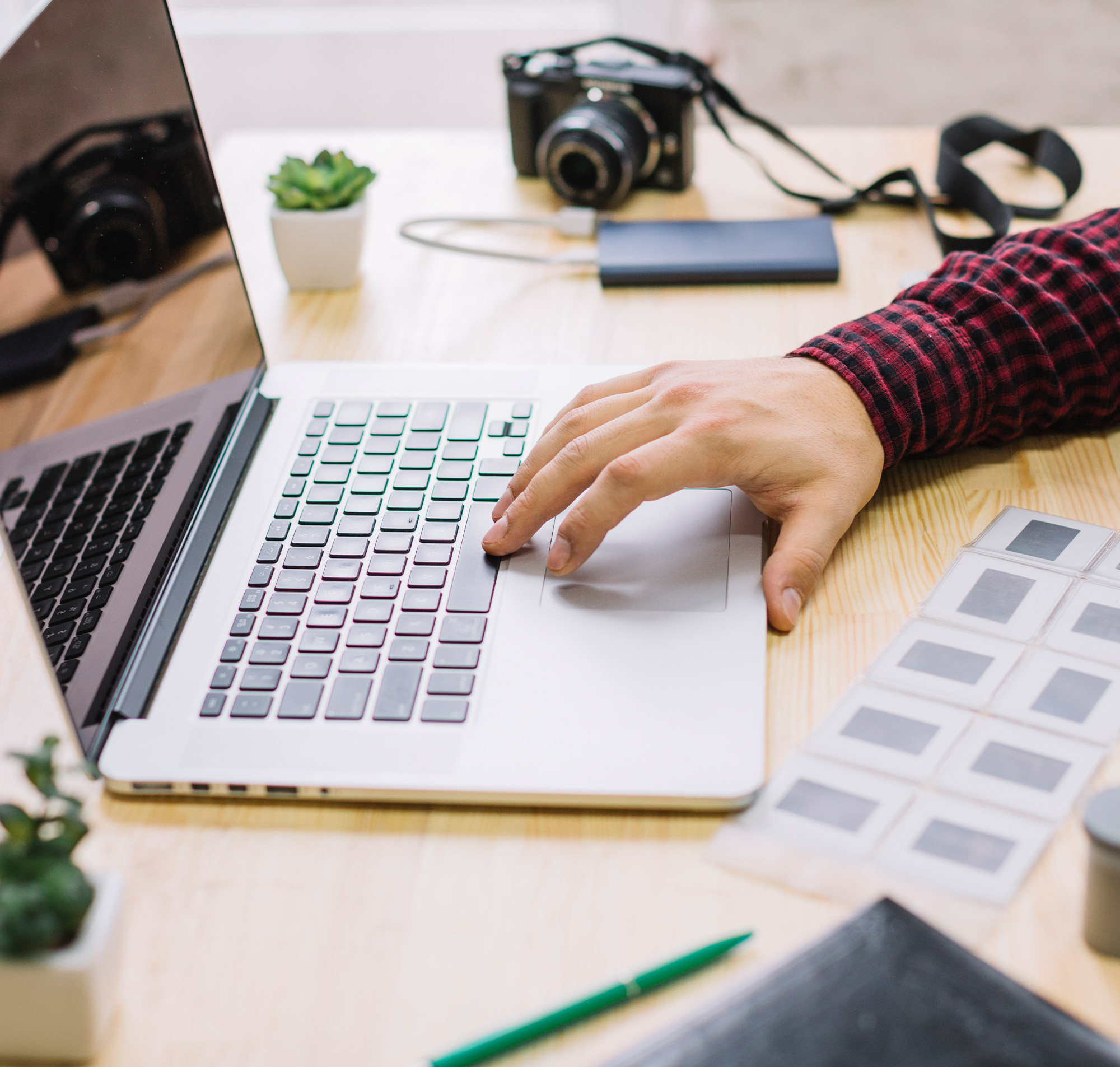Ein Mann arbeitet an einem Laptop, umgeben von Kameras, Pflanzen und Notizen auf einem Holztisch.
