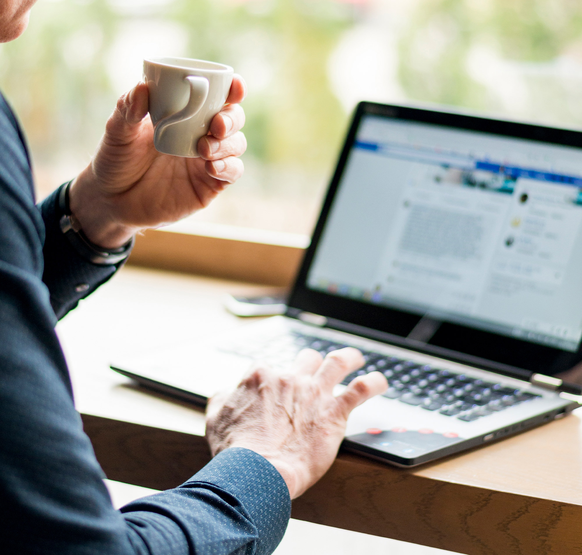 Ein Mann hält eine Tasse Kaffee und arbeitet an einem Laptop in einem modernen Café mit natürlichem Licht.