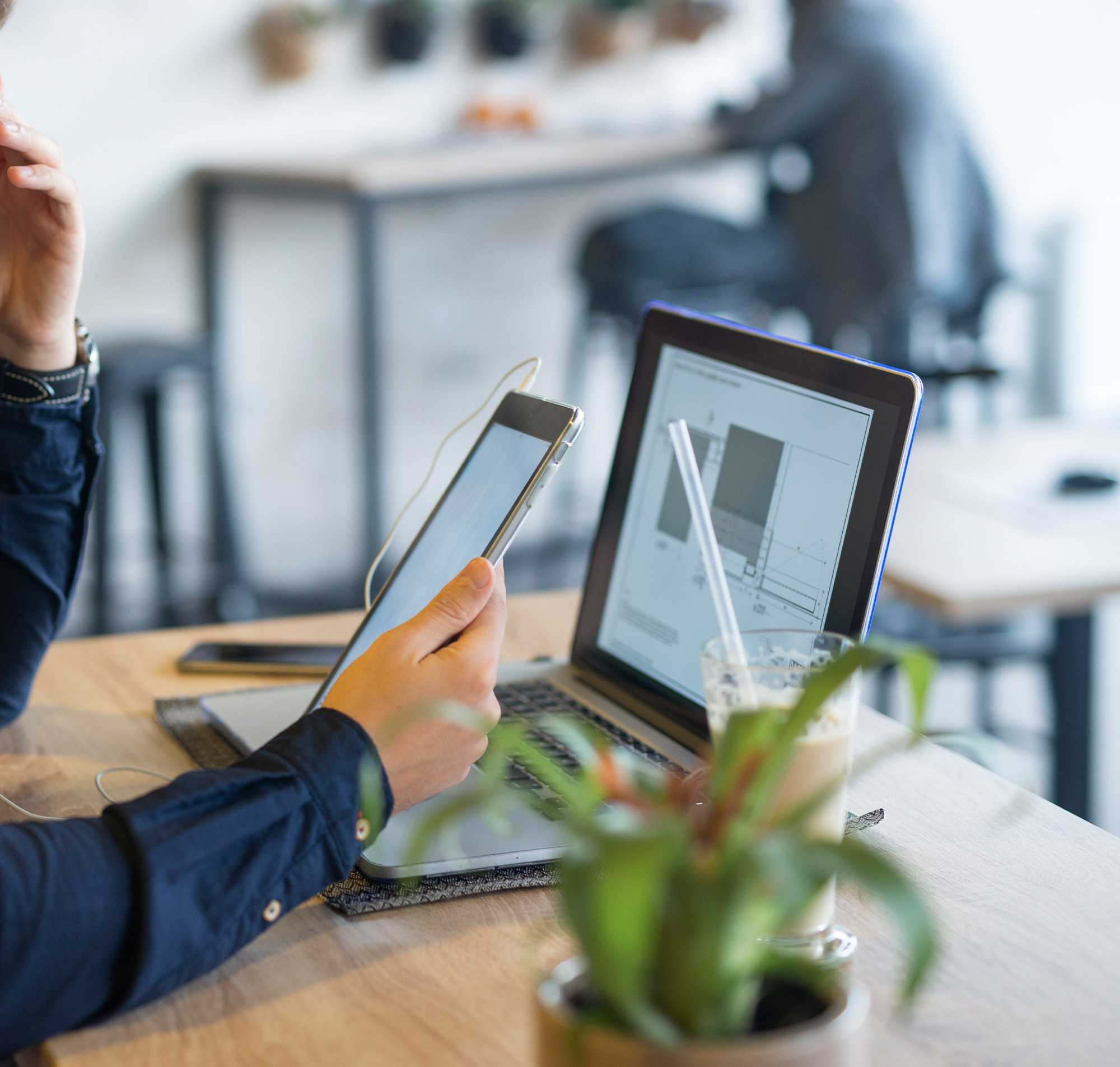 Eine Person arbeitet an einem Laptop und hält ein Tablet in einem modernen Café mit Pflanzen im Hintergrund.