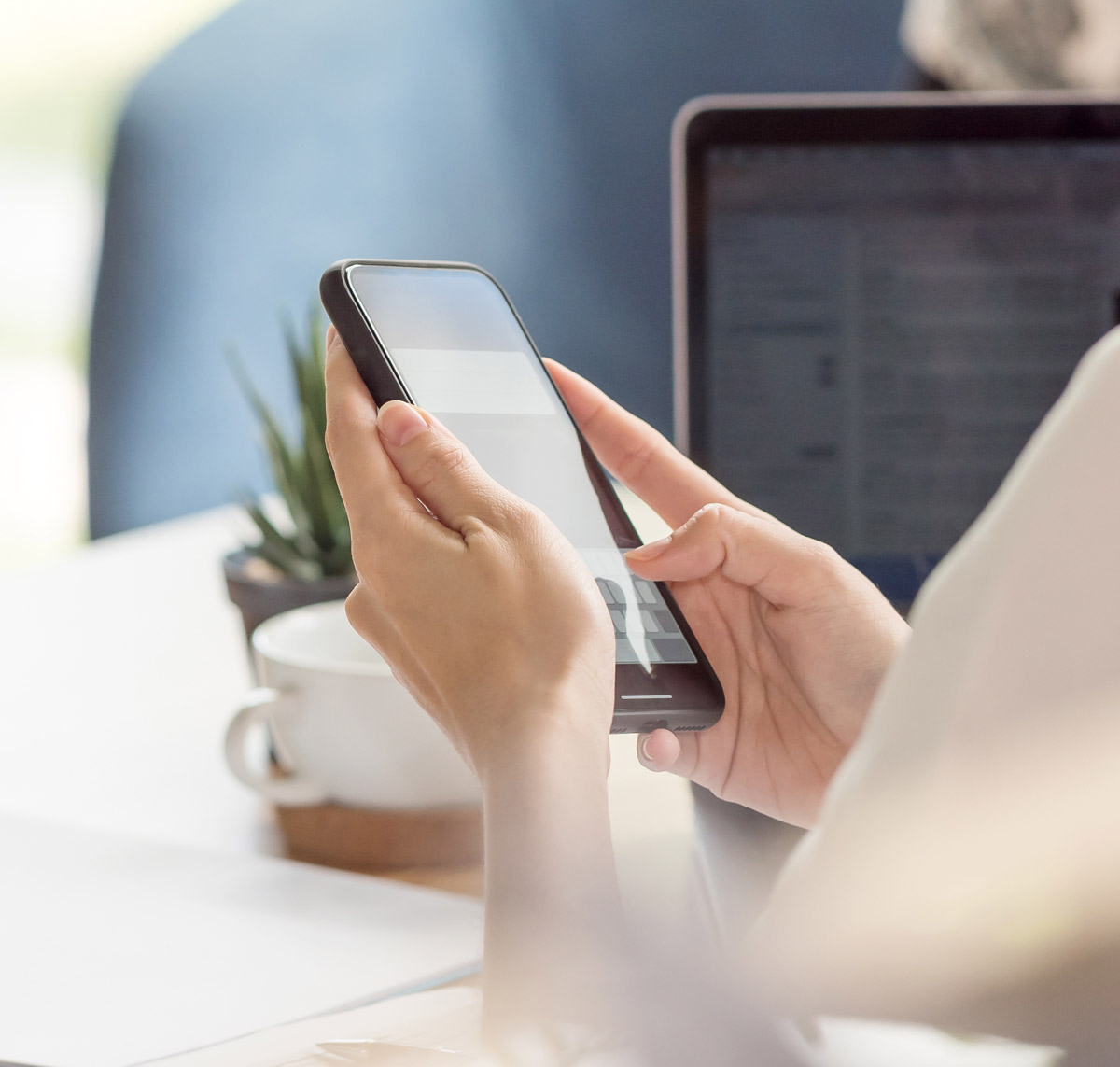 Eine Person hält ein Smartphone in der Hand und nutzt es, während ein Laptop im Hintergrund sichtbar ist.