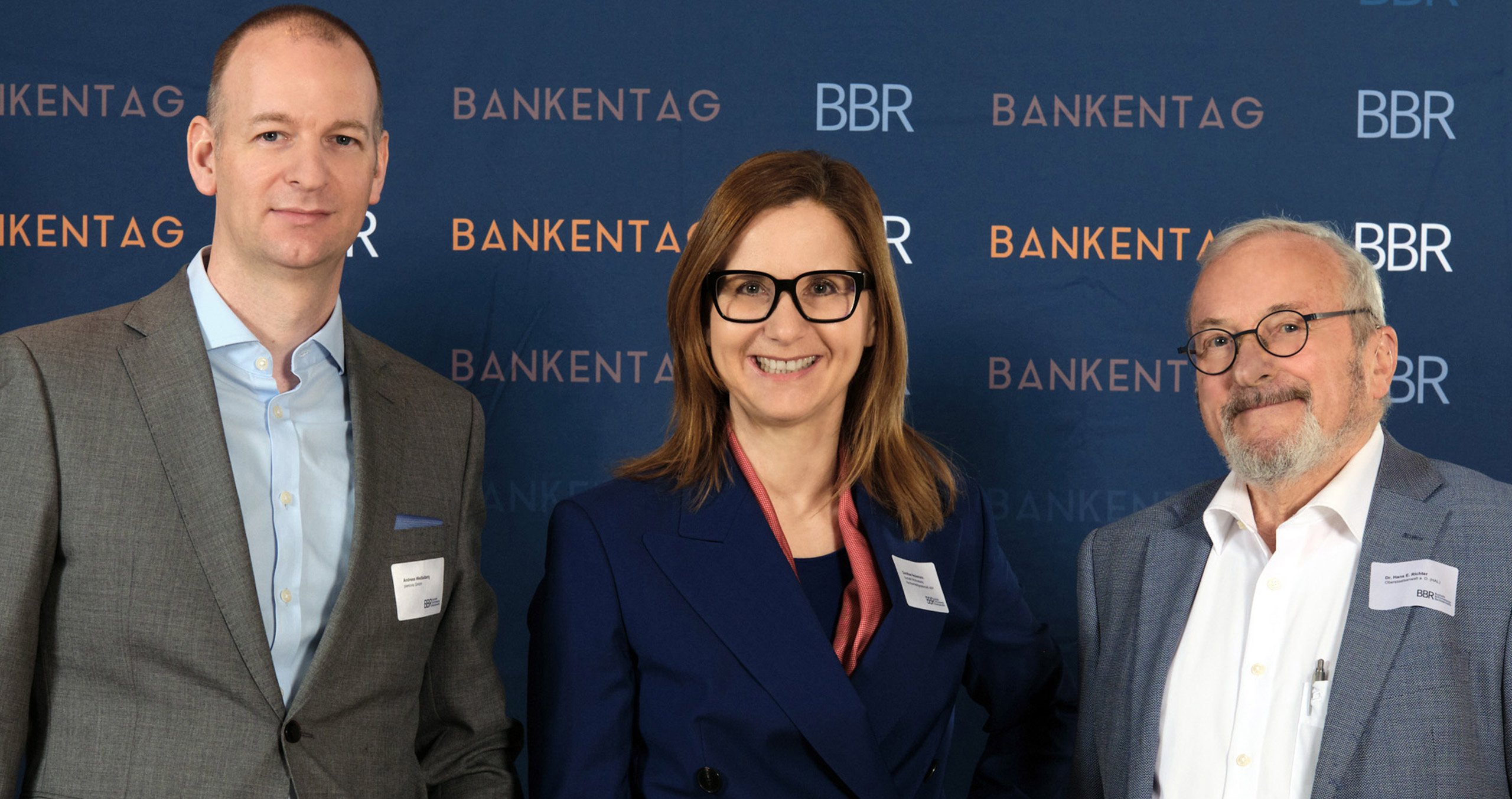 Drei Fachleute stehen beim Bankentag vor einem blauen Hintergrund mit dem Logo der Veranstaltung.