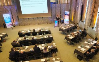 Konferenzraum mit Teilnehmern, die einem Vortrag auf einer großen Leinwand folgen, während sie an Tischen sitzen.