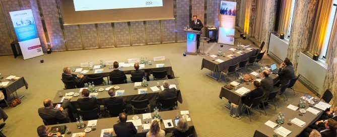 Konferenzraum mit Teilnehmern, die einem Vortrag auf einer großen Leinwand folgen, während sie an Tischen sitzen.
