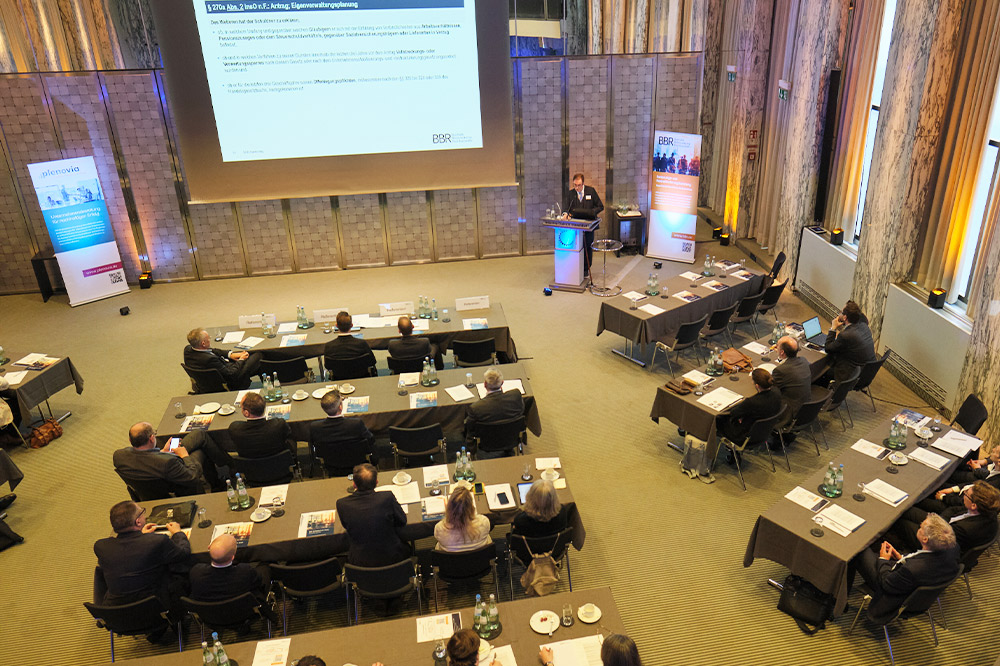 Konferenzraum mit Teilnehmern, die einem Vortrag auf einer großen Leinwand folgen, während sie an Tischen sitzen.