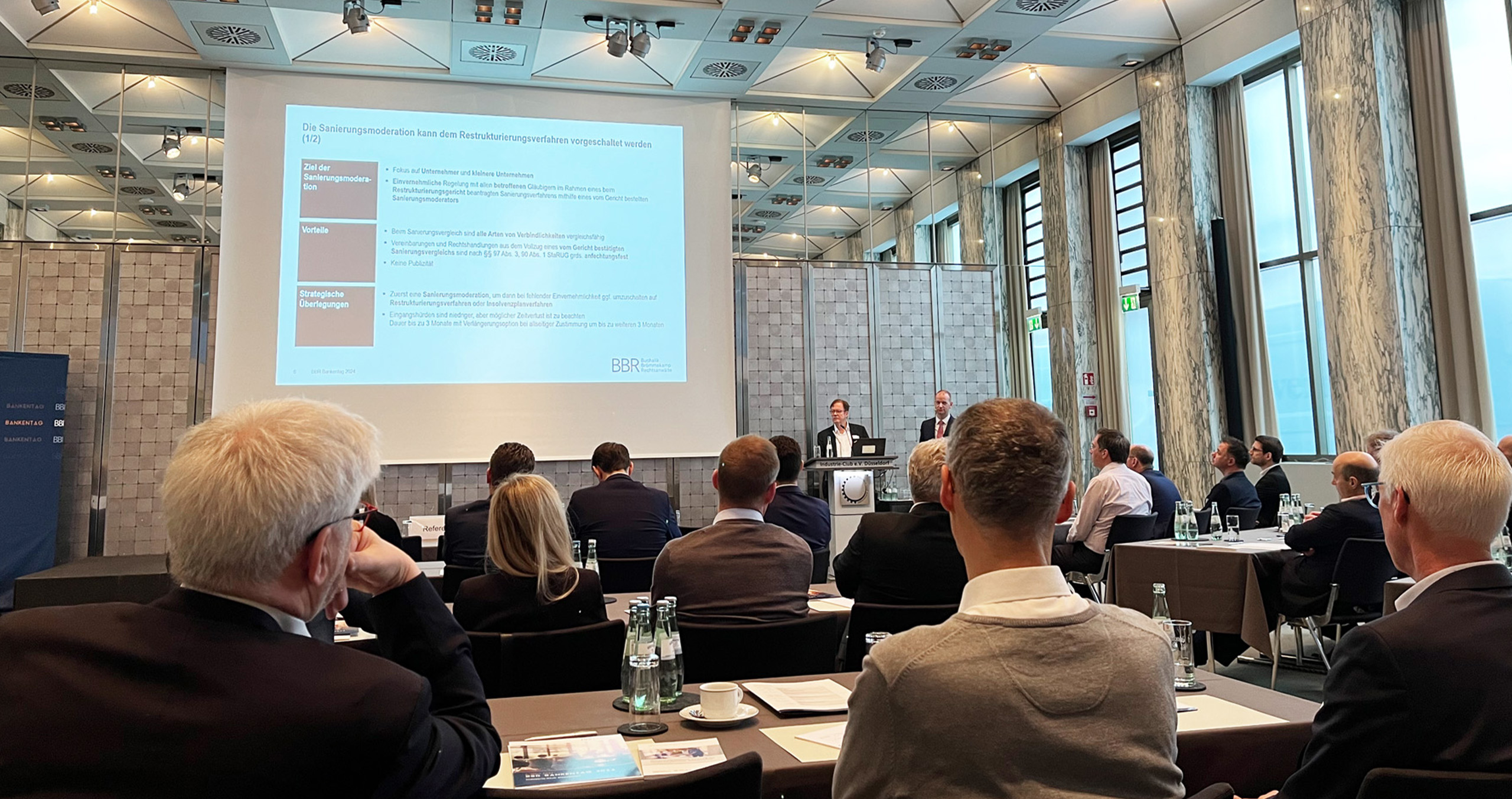 Teilnehmer eines Seminars zur Sanierungsmoderation in einem modernen Konferenzraum mit Präsentation im Hintergrund.