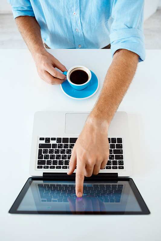 Eine Person in einem hellblauen Hemd sitzt an einem weißen Tisch, hält eine blaue Kaffeetasse und tippt mit dem Finger auf den Laptop-Bildschirm.