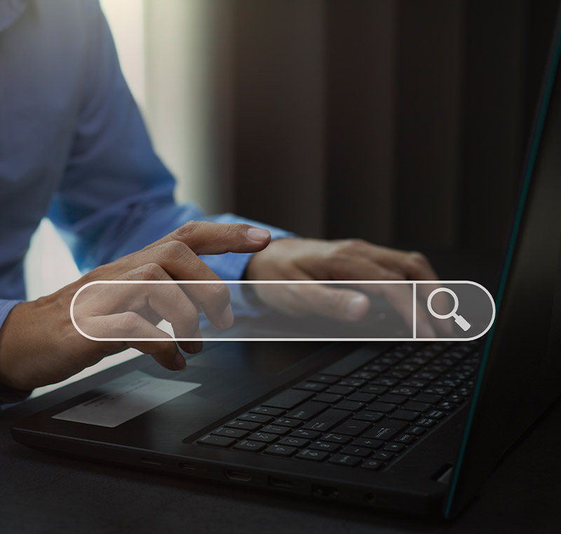 Eine Person tippt auf einer Laptop-Tastatur, während eine digitale Suchleiste mit einem Lupensymbol über dem Gerät eingeblendet ist.
