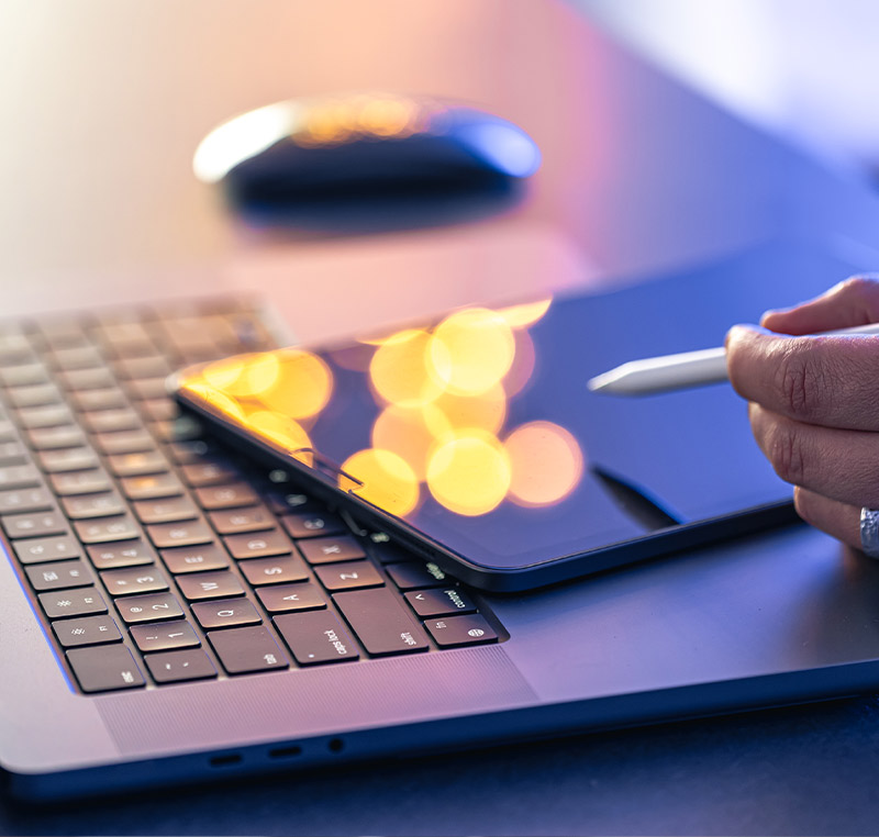 Eine Hand hält einen Eingabestift über ein Tablet, das auf einer Laptop-Tastatur liegt, mit unscharfen Lichtreflexen im Hintergrund.